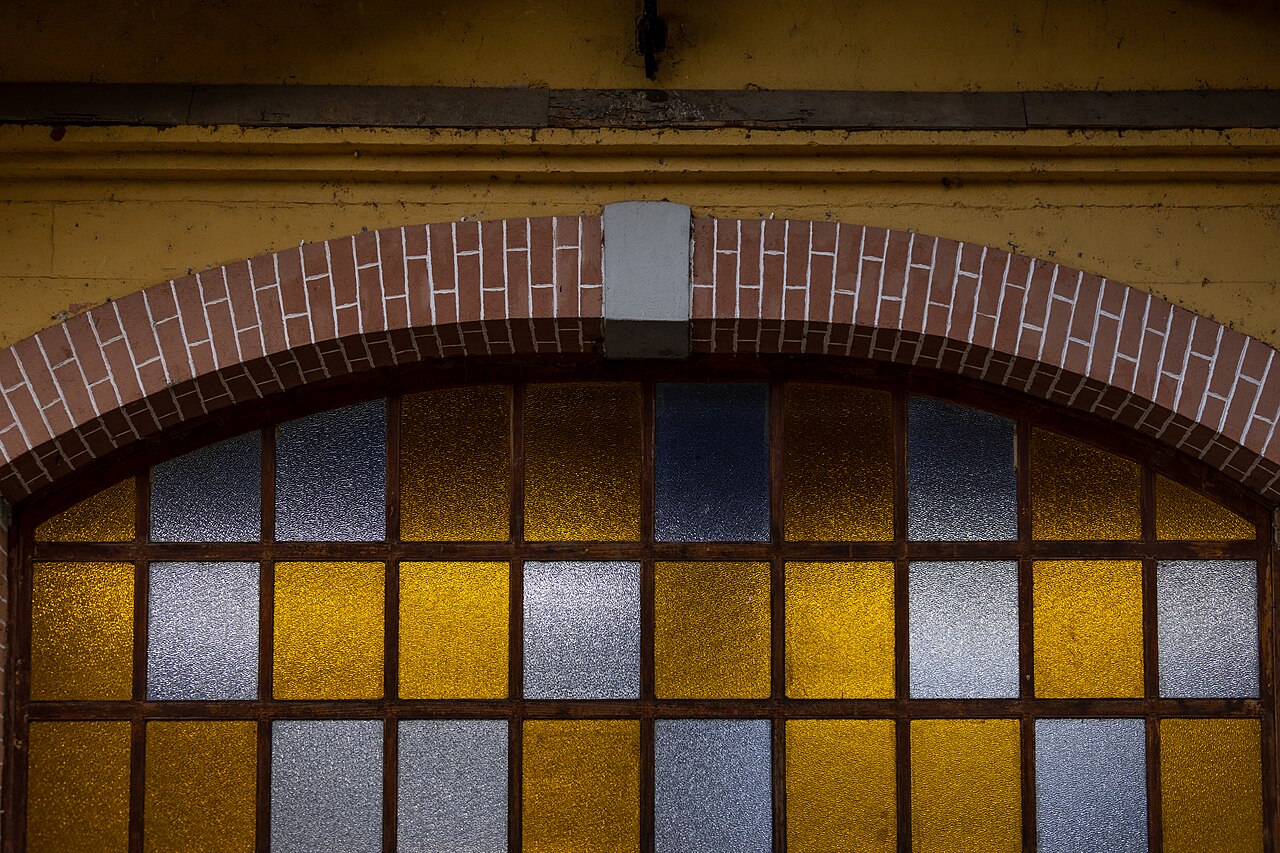 Janela arqueada com caixilhos escuros e vidros amarelos e translúcidos, sob arco de tijolos em parede amarela desgastada.