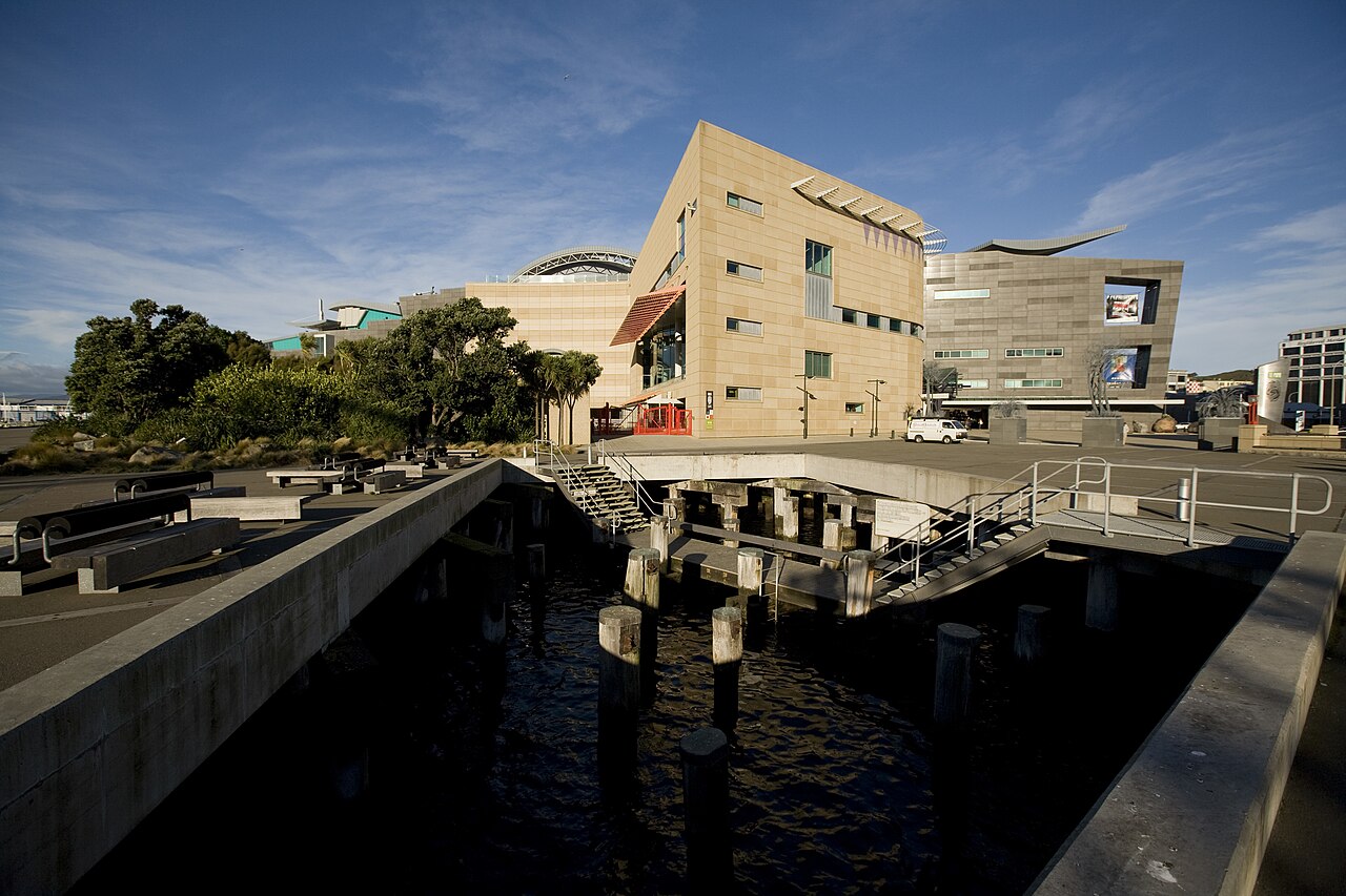 Edifício contemporâneo do Te Papa junto à orla, com água, estruturas de concreto, estacas de madeira e corrimãos em primeiro plano.