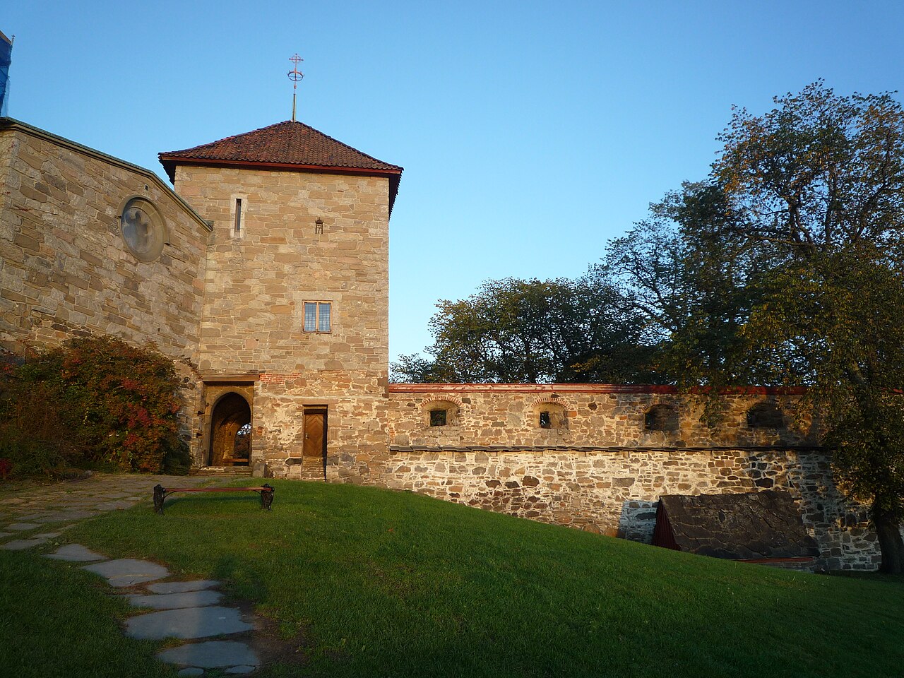 Construções de pedra de Akershus com torre de telhado avermelhado, passagem em arco, gramado e caminho de lajes.