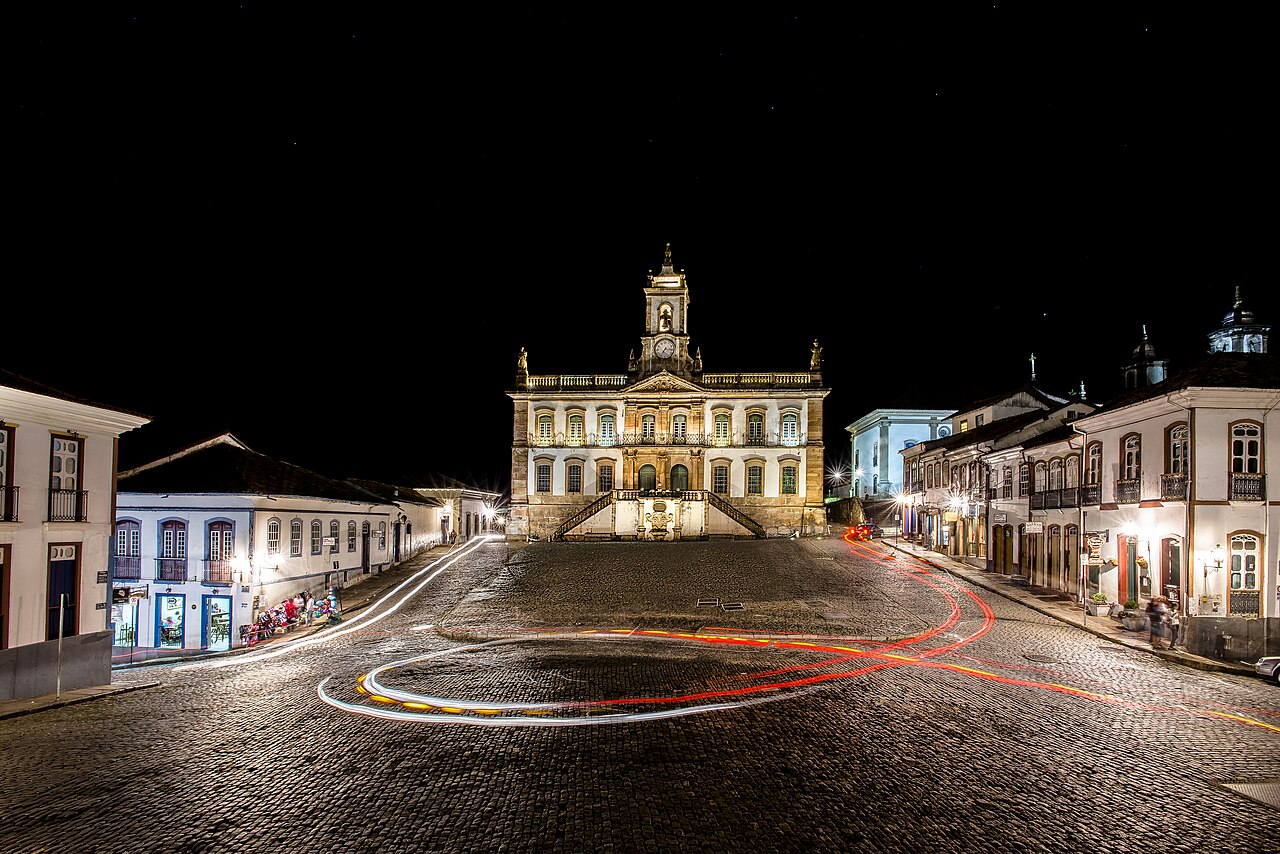 Vista noturna do Museu da Inconfidência iluminado, com torre de relógio, escadarias simétricas e rastros luminosos de veículos na praça.