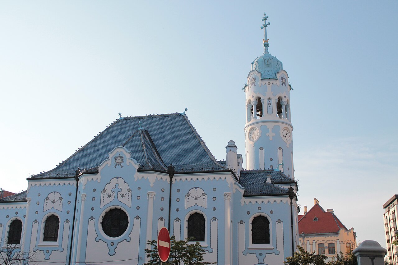 Igreja azul-clara com ornamentos brancos, telhado azul e torre com relógios e cruz, com vegetação e uma placa à frente.