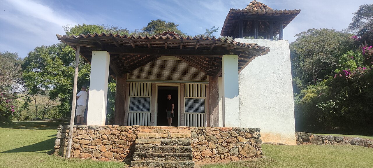 Fachada branca da Capela do Sítio Santo Antônio com alpendre de madeira, torre lateral, degraus de pedra e duas pessoas junto à entrada.