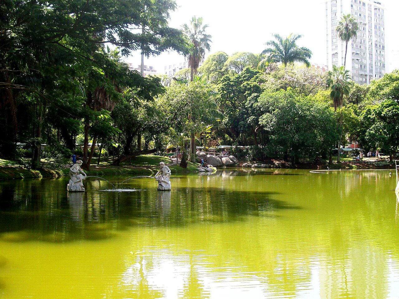 Lago do Campo de São Bento com esculturas brancas, jatos de fonte, árvores e palmeiras, com edifícios ao fundo.