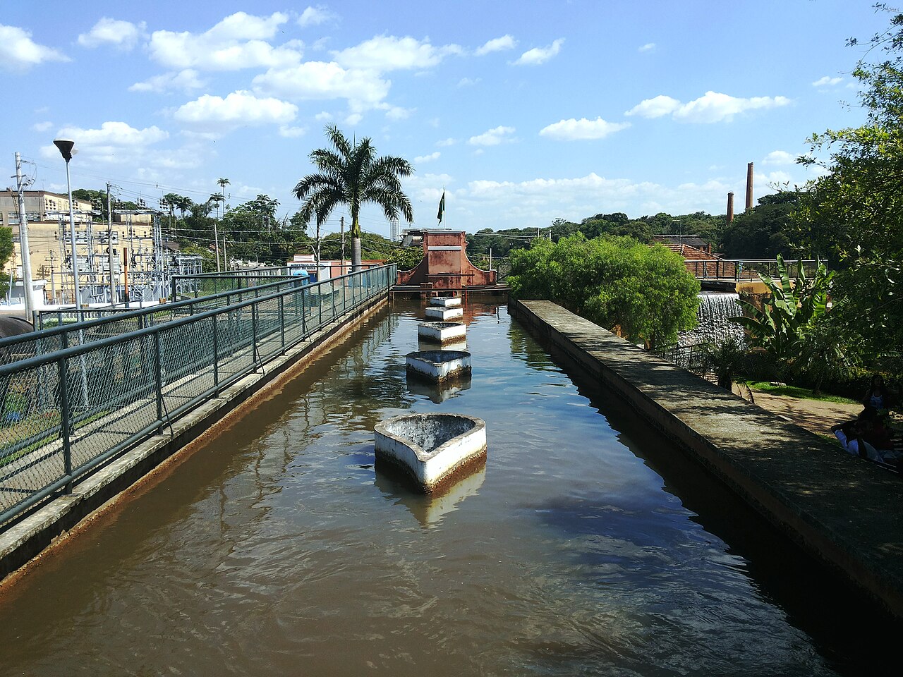 Canal com água e estruturas brancas alinhadas, passarela com grades à esquerda e instalações ao fundo no Museu da Água.