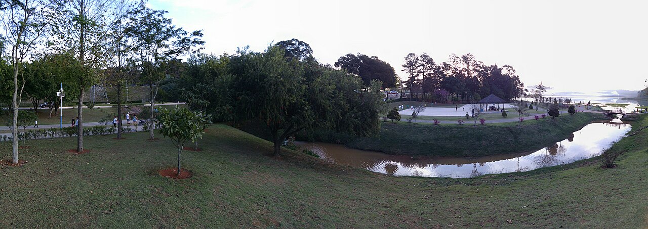 Panorâmica de parque com gramados, árvores, caminhos com pessoas, um pavilhão e um canal de água curvo.