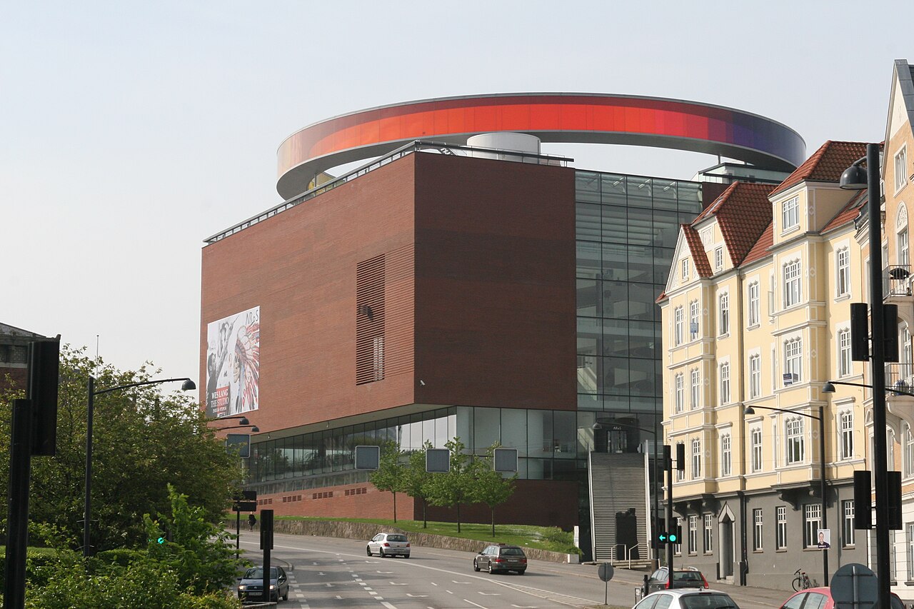 Edifício de tijolos avermelhados do ARoS com passarela circular de painéis coloridos no topo, árvores e uma rua com carros à frente.