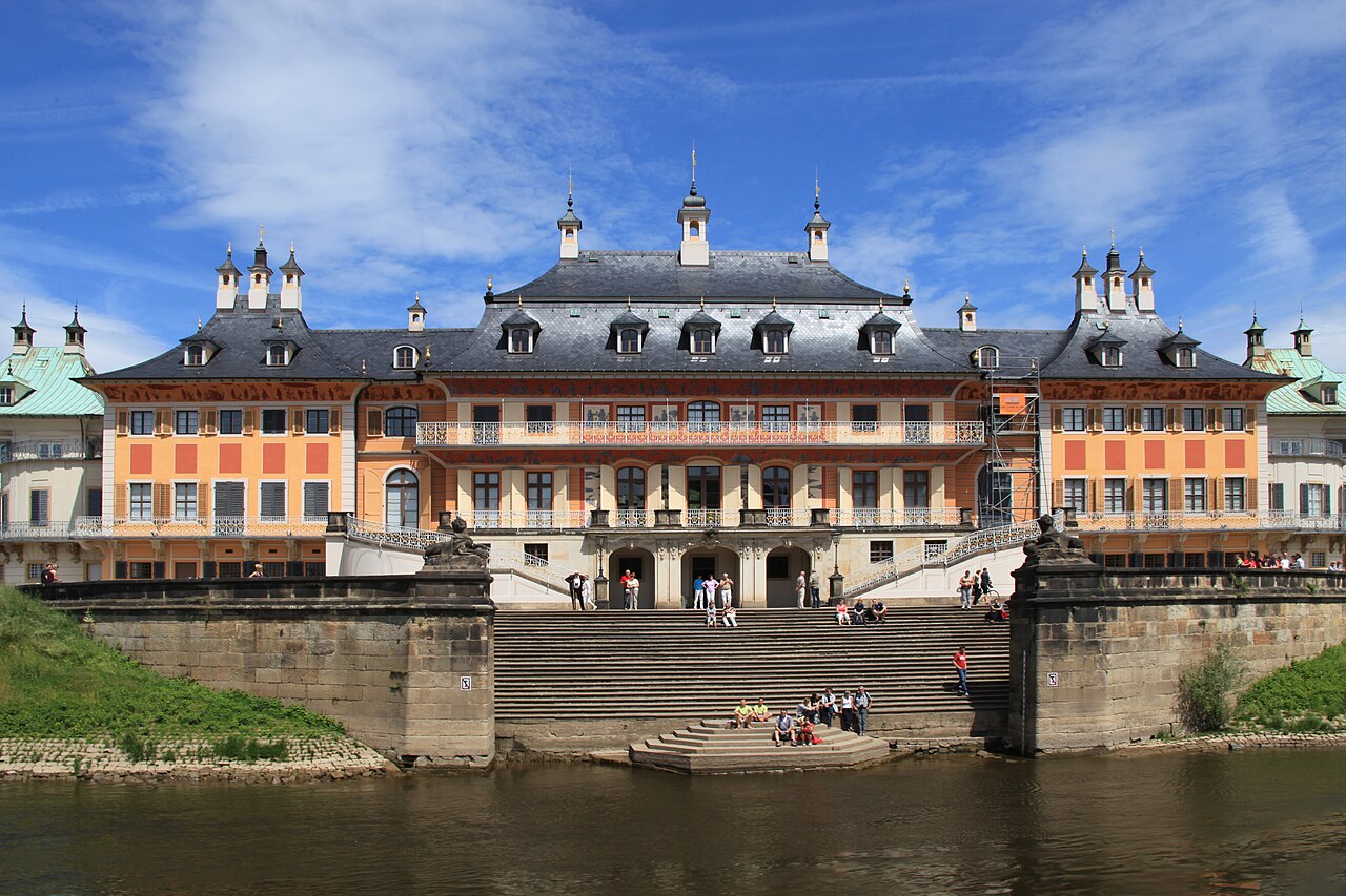 Palácio de Pillnitz visto do rio, com fachada amarela e salmão, escadaria até a água e andaimes à direita.