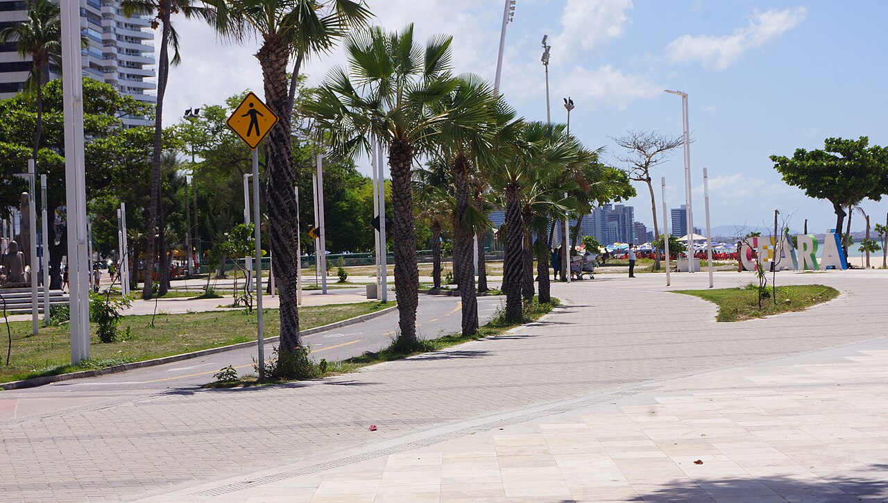 Calçadão da orla de Fortaleza com palmeiras, ciclovia, edifícios, pessoas e uma faixa de mar ao fundo.