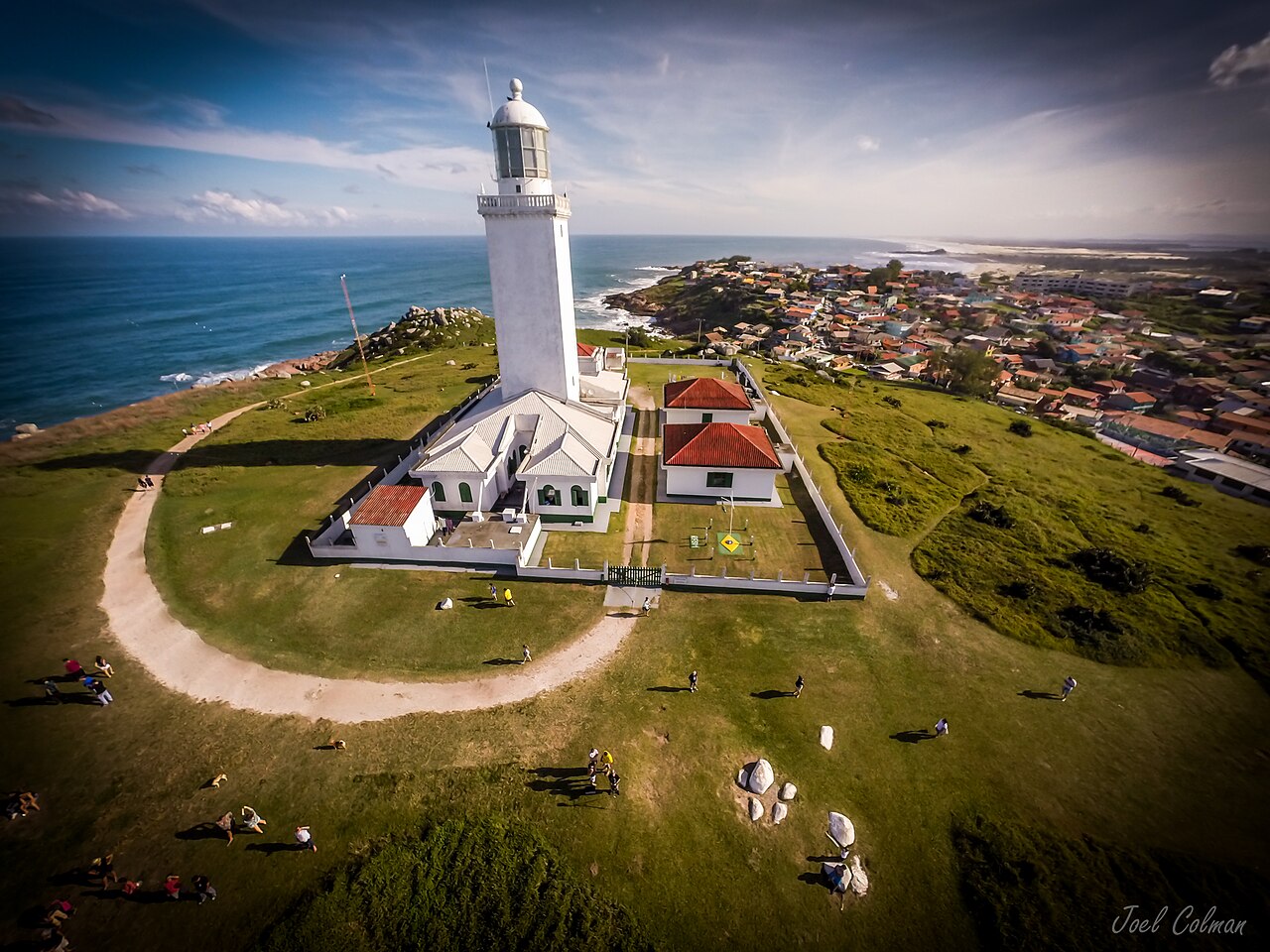 Vista elevada de um farol branco em área gramada e murada, com edifícios anexos, caminhos, costão rochoso, mar e povoado com praias ao fundo.