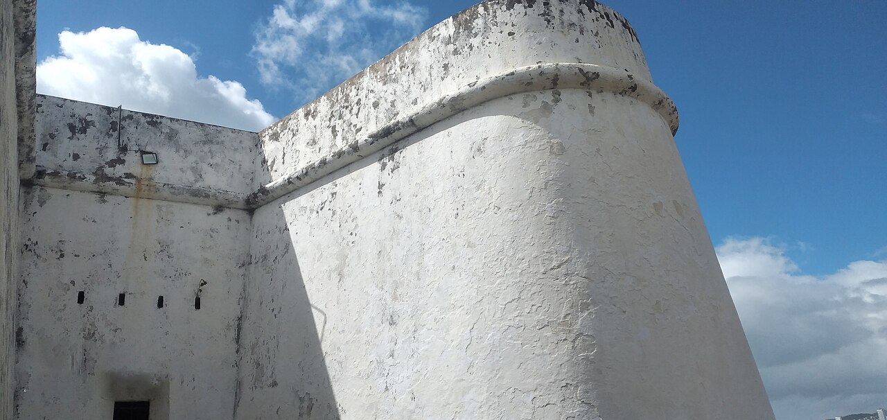 Muralhas brancas do Forte dos Reis Magos, com trecho arredondado, pequenas aberturas e pintura descascada, sob céu azul com nuvens.