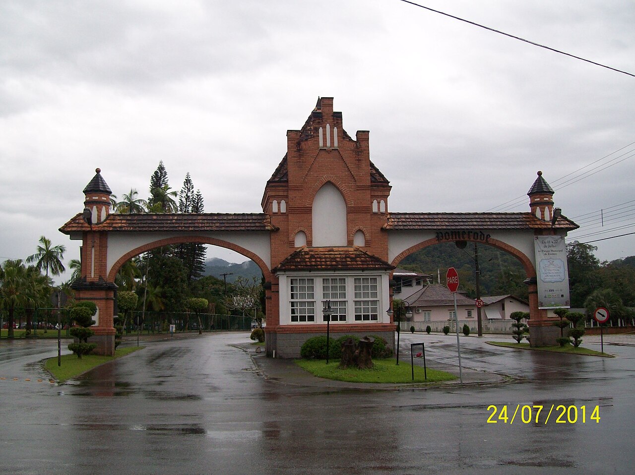 Portal Norte de tijolos com dois arcos, telhados e pequenas torres, sob céu encoberto e diante de pavimento molhado.