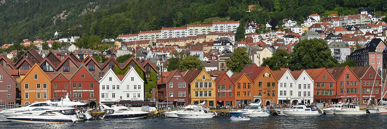 Fachadas estreitas e coloridas de Bryggen diante do cais, com barcos em primeiro plano e uma encosta com casas ao fundo.