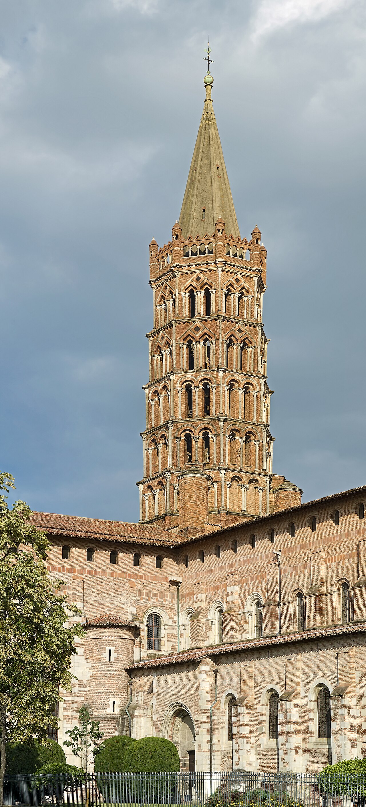 Exterior de Saint-Sernin em tijolos e pedra, com torre octogonal de vários níveis, árvores e arbustos à frente.