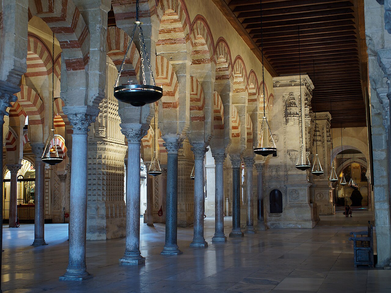 Fileiras de colunas e arcos com faixas avermelhadas e claras no interior da Mezquita-Catedral, com luminárias suspensas e teto de madeira.