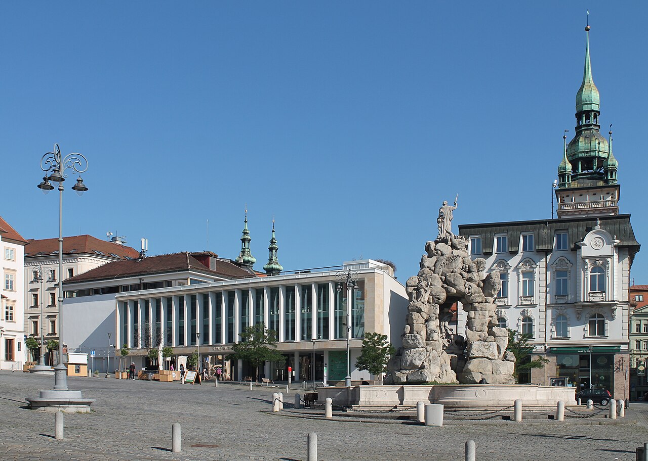 Fonte monumental com esculturas no centro de uma praça de paralelepípedos, cercada por fachadas históricas e um edifício moderno em Zelný trh.