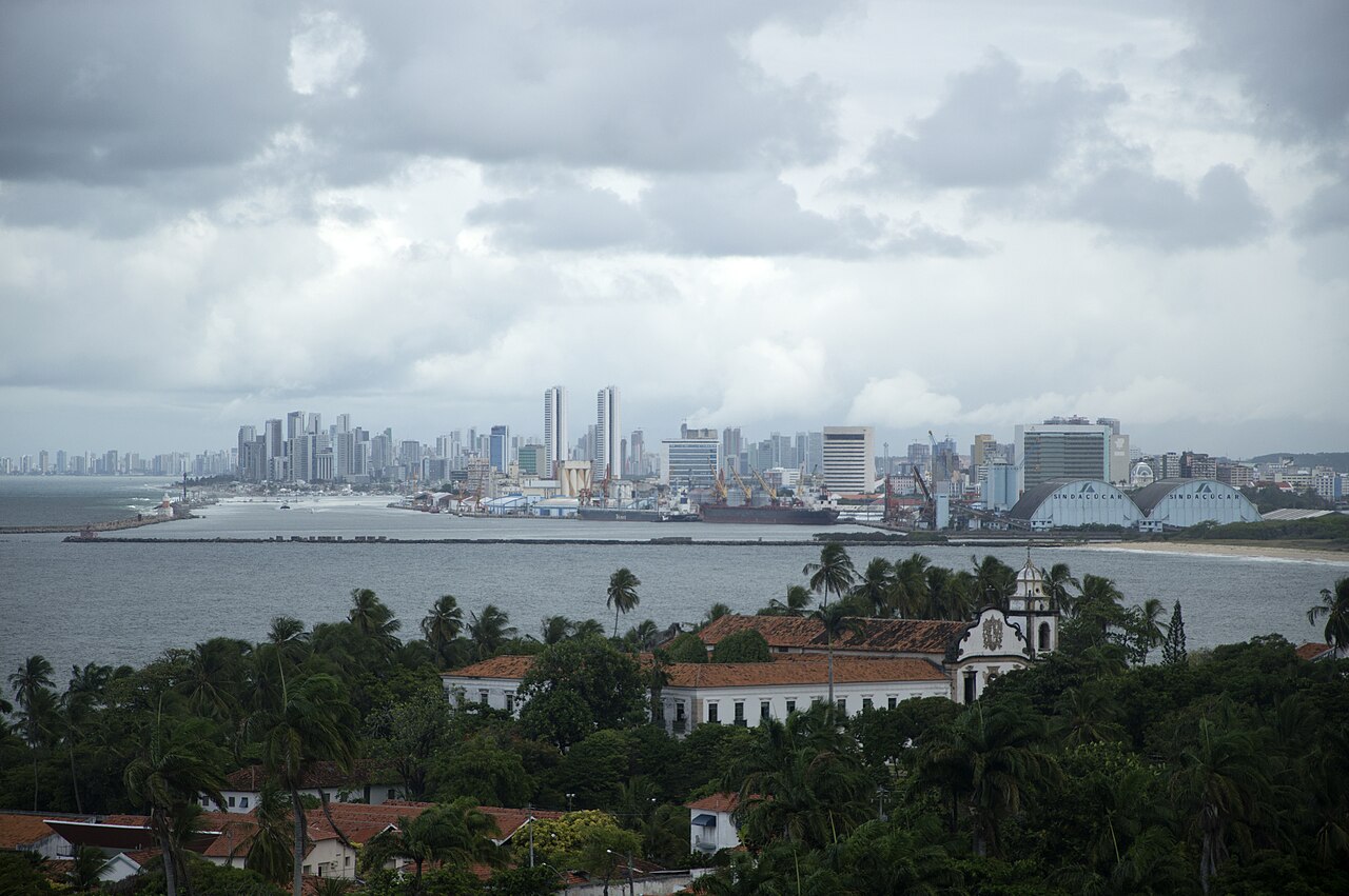 Conjunto de São Bento com paredes brancas, detalhes escuros, torre e telhados cerâmicos entre árvores, com água, guindastes e edifícios de Recife ao fundo.