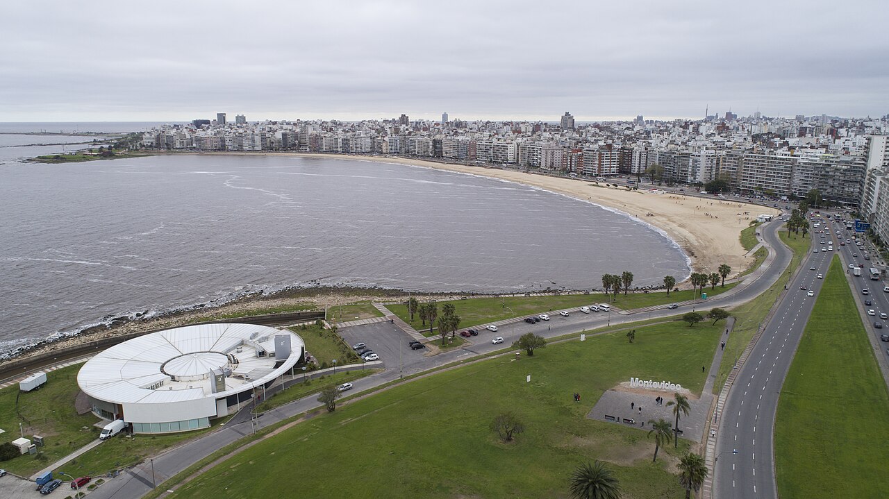 Vista elevada da praia curva de Pocitos sob céu encoberto, com edifícios na orla e letreiro de Montevideo entre gramados e avenidas.