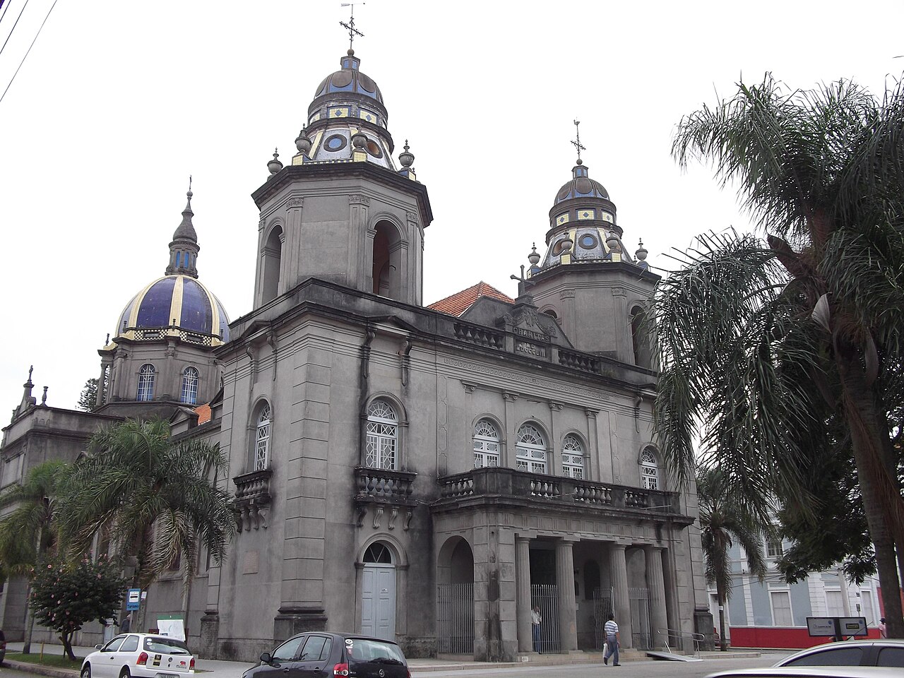 Fachada cinza da Catedral São Francisco de Paula, com duas torres, cúpulas com detalhes azuis e amarelos, palmeiras, carros e pedestres à frente.