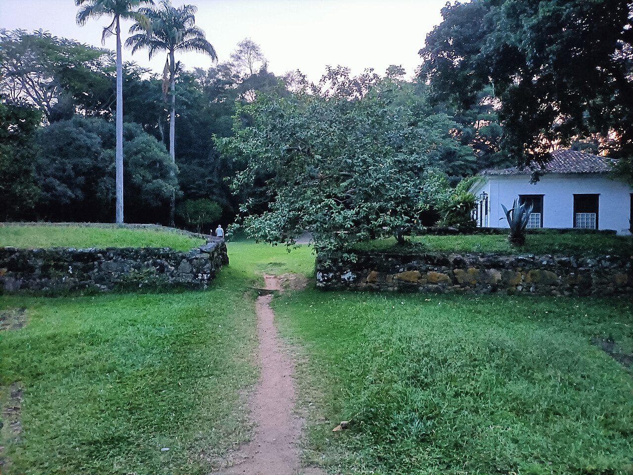 Caminho de terra entre gramados e muretas de pedra no Forte Defensor Perpétuo, com árvores e uma construção branca à direita.