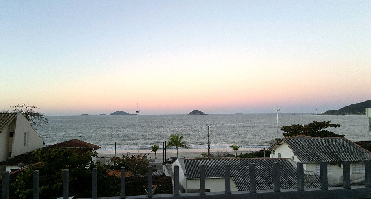 Vista elevada da orla com casas, palmeiras, rua, faixa de areia e mar com pequenas ilhas sob céu de tons suaves.