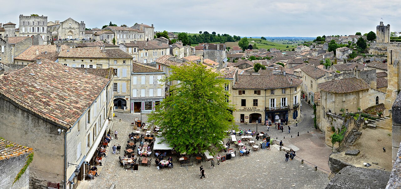 Vista elevada de uma praça com mesas, pessoas e árvore central em Saint-Émilion, cercada por construções de pedra e telhados cerâmicos.