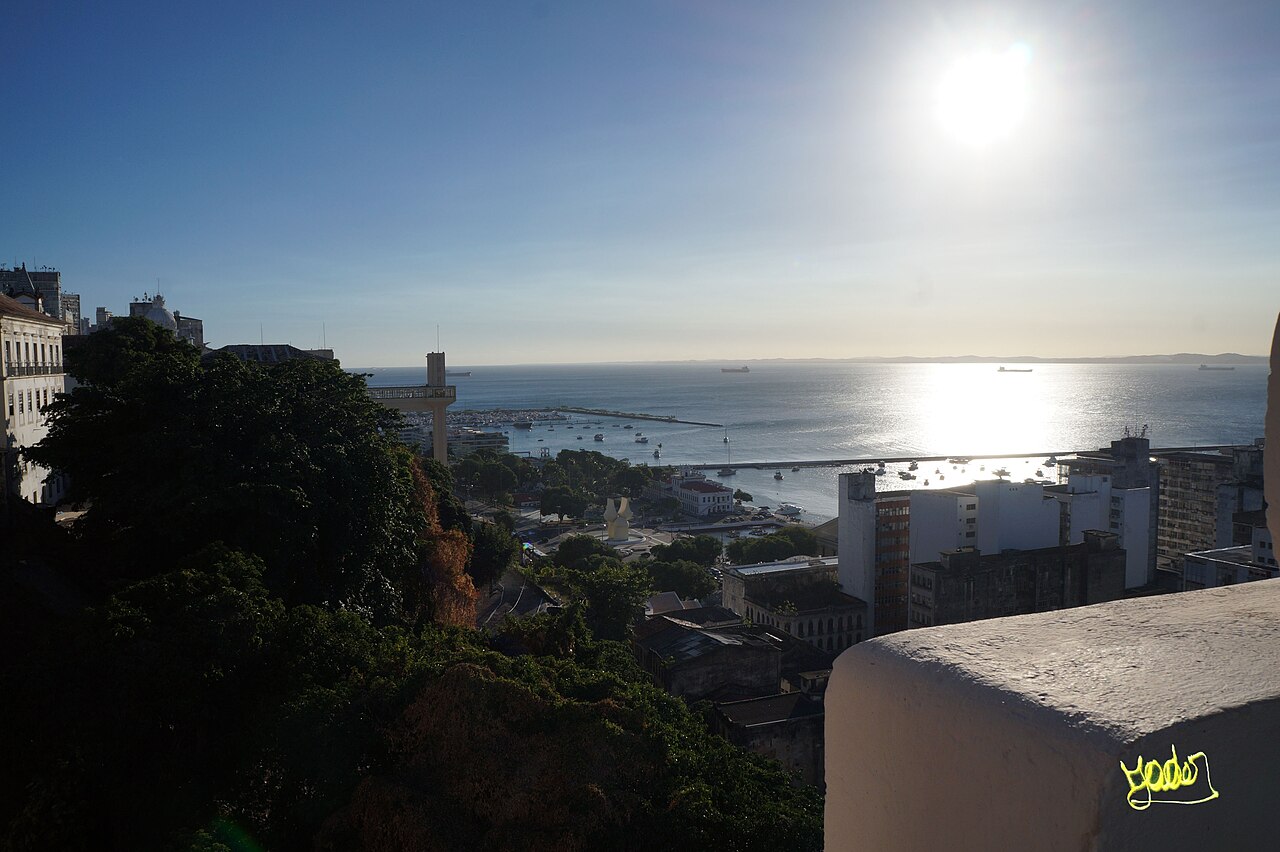 Vista elevada da baía com barcos e edifícios; a torre e a passarela do Elevador Lacerda aparecem parcialmente encobertas por árvores.