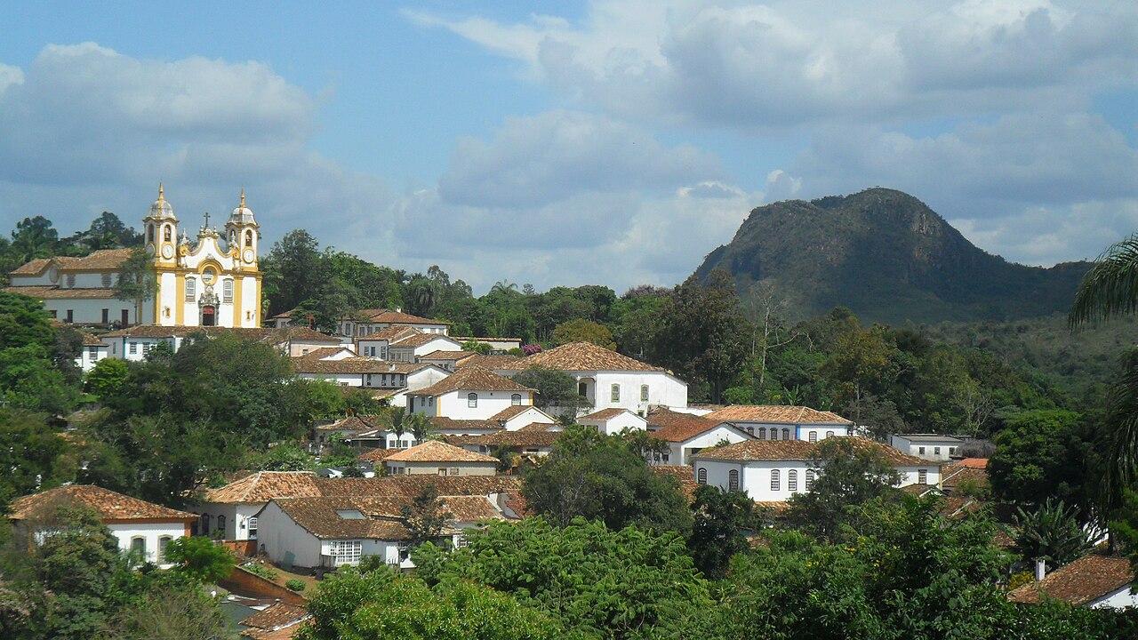 Vista panorâmica da Matriz de Santo Antônio, branca e amarela, com duas torres, casario de telhados cerâmicos, vegetação e elevação rochosa ao fundo.