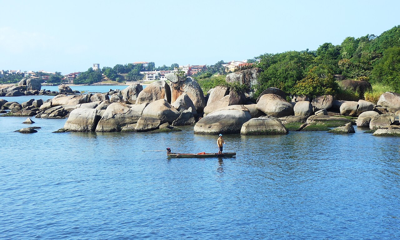 Água azul com pequenas ondulações, pessoa em pé numa embarcação estreita, rochas junto à margem, vegetação e construções ao fundo.