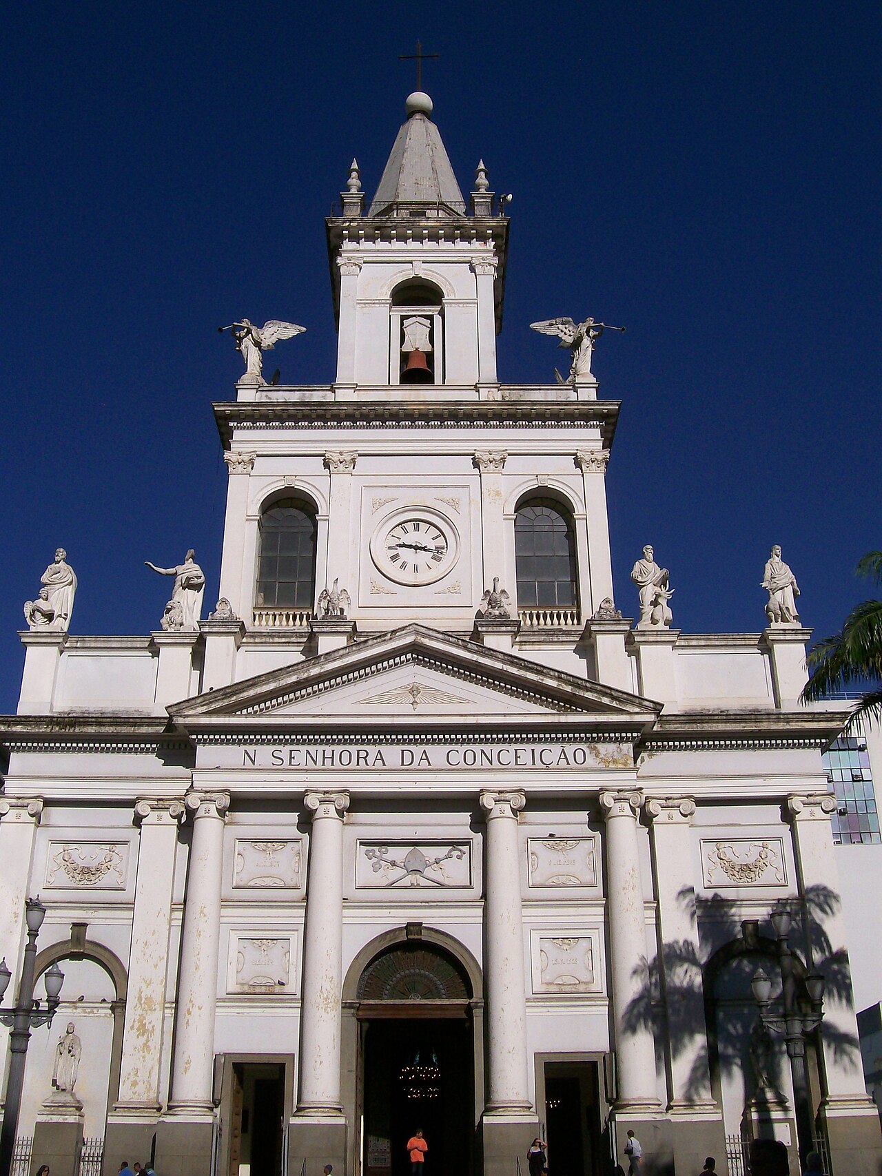 Fachada clara da Catedral Metropolitana de Campinas, com colunas, frontão triangular, relógio e torre central com sino e cruz.