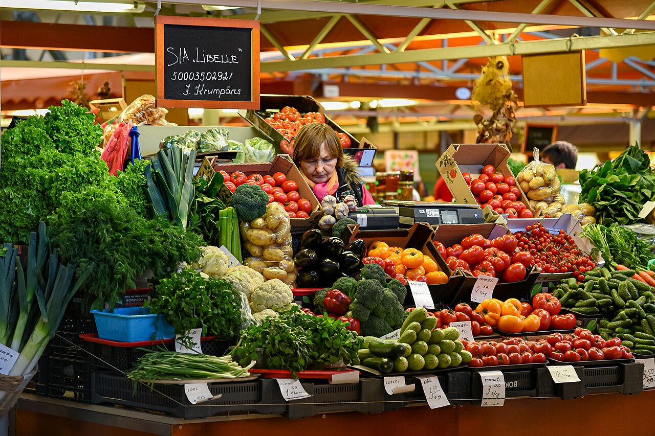 Banca coberta do Mercado Central de Riga com tomates, folhas, brócolis, couves-flores e uma vendedora atrás do balcão.
