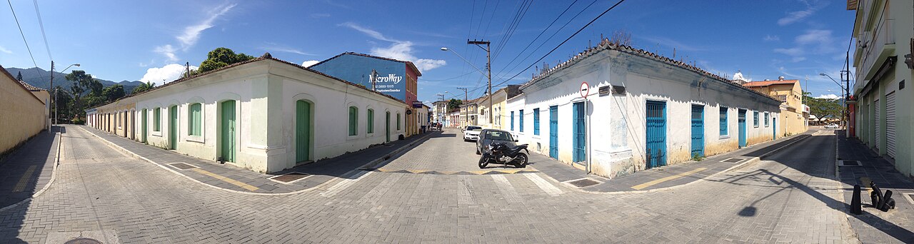 Cruzamento no Centro de São Sebastião com casario térreo claro, portas e janelas verdes e azuis, veículos, fiação e morros ao fundo.