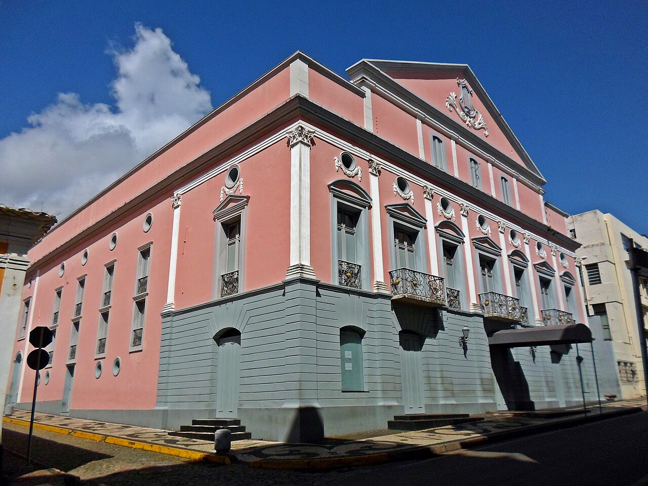 Fachadas rosa e cinza do Teatro Arthur Azevedo em uma esquina, com pilastras brancas, sacadas ornamentadas e uma lira no frontão.