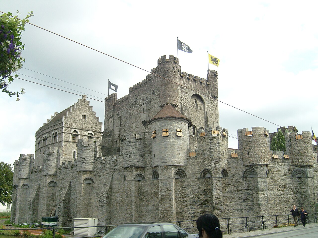 Exterior do Gravensteen, com muralhas de pedra ameadas, torres e bandeiras; fios aéreos, um automóvel e pedestres aparecem à frente.