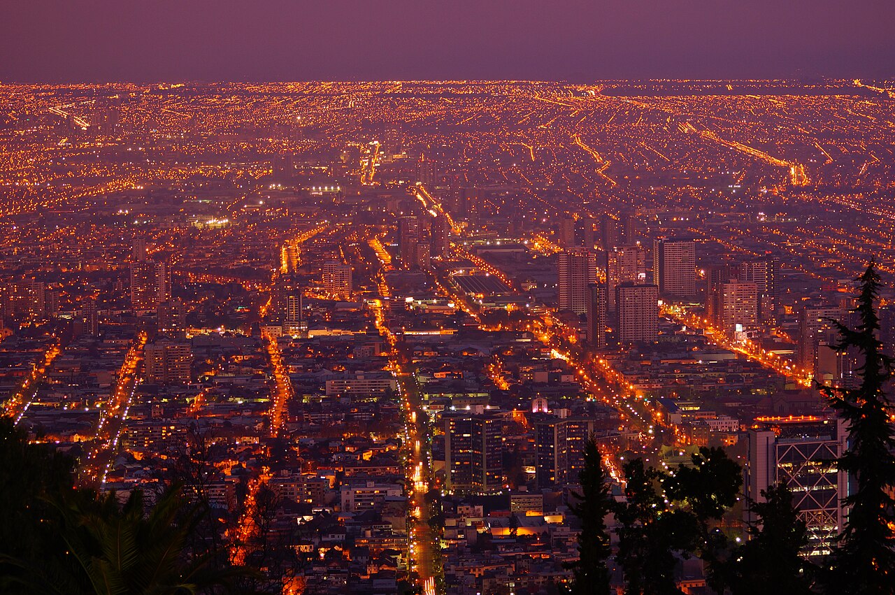 Panorama urbano ao anoitecer com ruas iluminadas, edifícios, céu arroxeado e árvores escuras em primeiro plano.