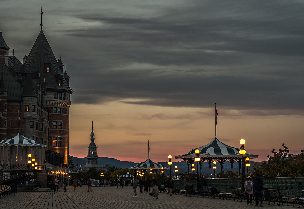 Calçadão de madeira da Terrasse Dufferin com pedestres, bancos, postes acesos e quiosques listrados, junto a parte do Château Frontenac, sob céu nublado com luz alaranjada no horizonte.