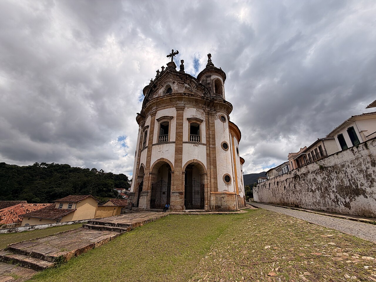 Exterior da Igreja de Nossa Senhora do Rosário, com fachada curva, elementos de pedra, degraus e gramado sob céu nublado.