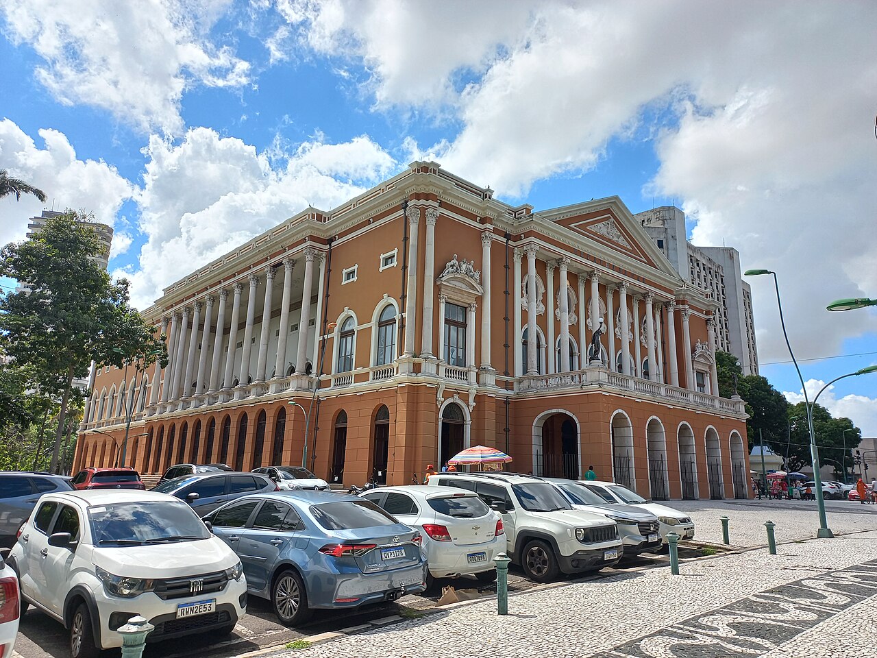 Fachada do Theatro da Paz em tons de terracota, com colunas claras, frontão triangular, esculturas e árvores ao redor.