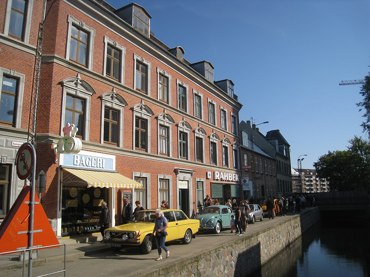 Rua do setor de 1974 de Den Gamle By junto a um canal, com prédios de tijolos, comércio com letreiro BAGERI, pedestres e carros antigos.