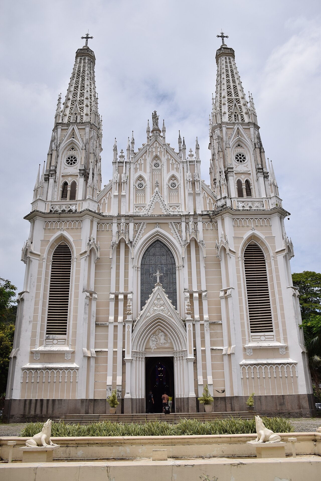 Fachada bege e branca da Catedral Metropolitana de Vitória, com duas torres, arcos ogivais, rosáceas e pessoas junto à entrada.