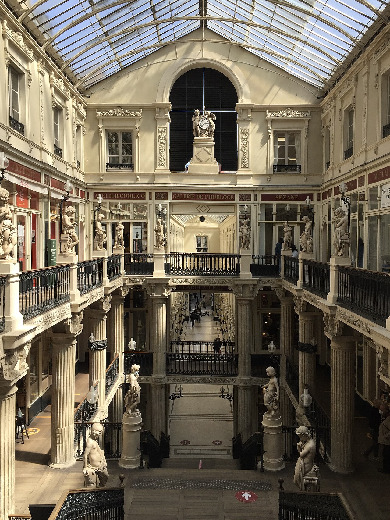 Interior do Passage Pommeraye com cobertura de vidro, escadarias entre níveis, colunas ornamentadas, estátuas e relógio central.