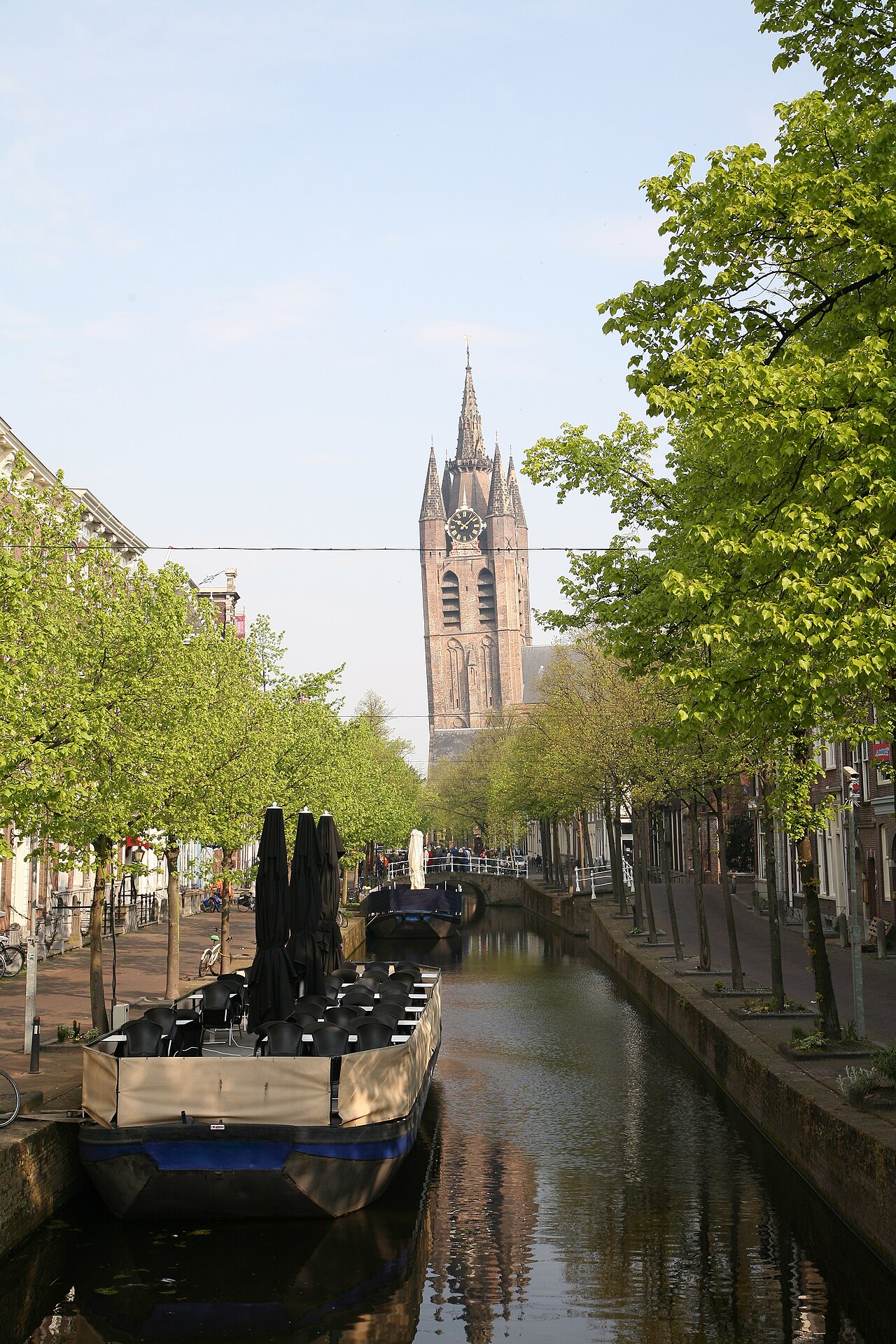 Canal arborizado com construções de tijolos, uma embarcação com mesas e ombrelones fechados e a torre da Oude Kerk ao fundo.