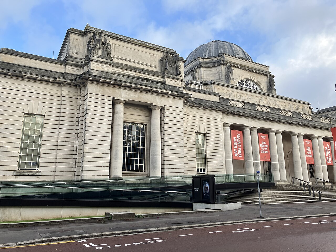 Fachada de pedra clara do National Museum Cardiff, com colunas, cúpula, escadaria e faixas vermelhas bilíngues.