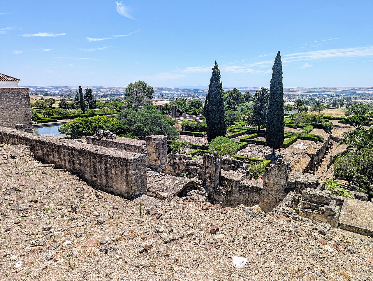 Vista elevada de Medina Azahara com ruínas de pedra, fragmentos de colunas, sebes geométricas, ciprestes e uma planície ao fundo.