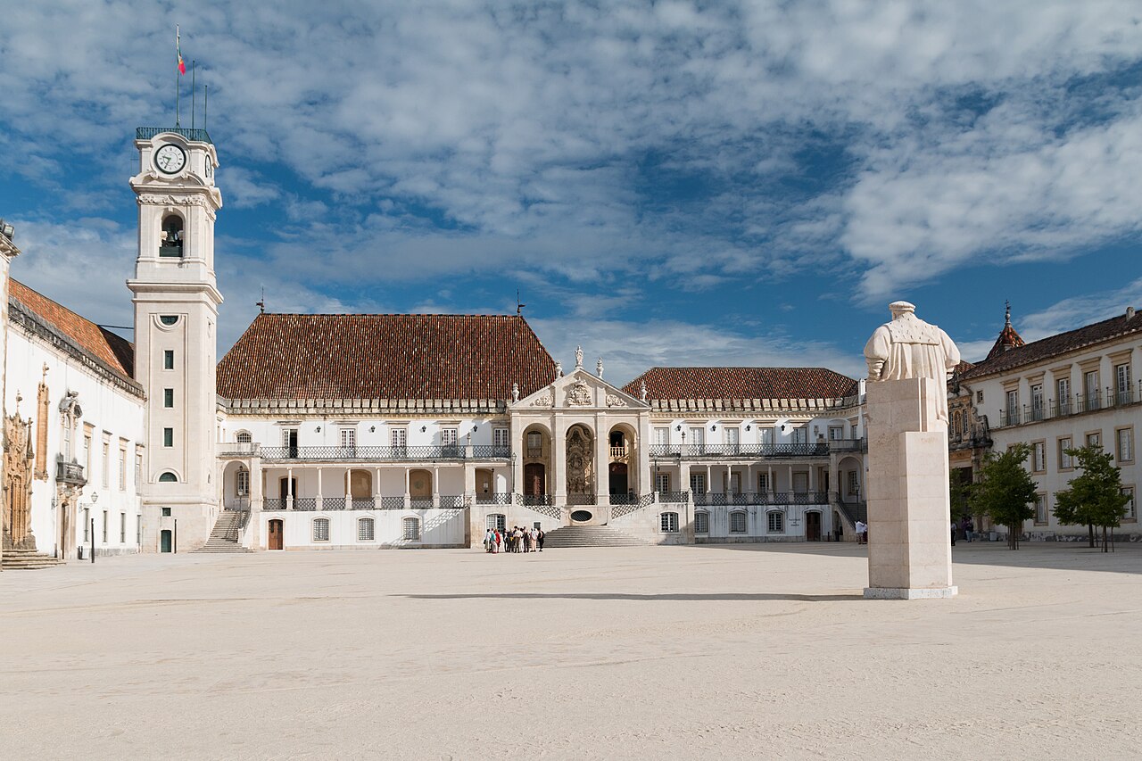 Pátio pavimentado do Paço das Escolas, cercado por edifícios claros, com torre de relógio à esquerda, escadarias e estátua à direita.