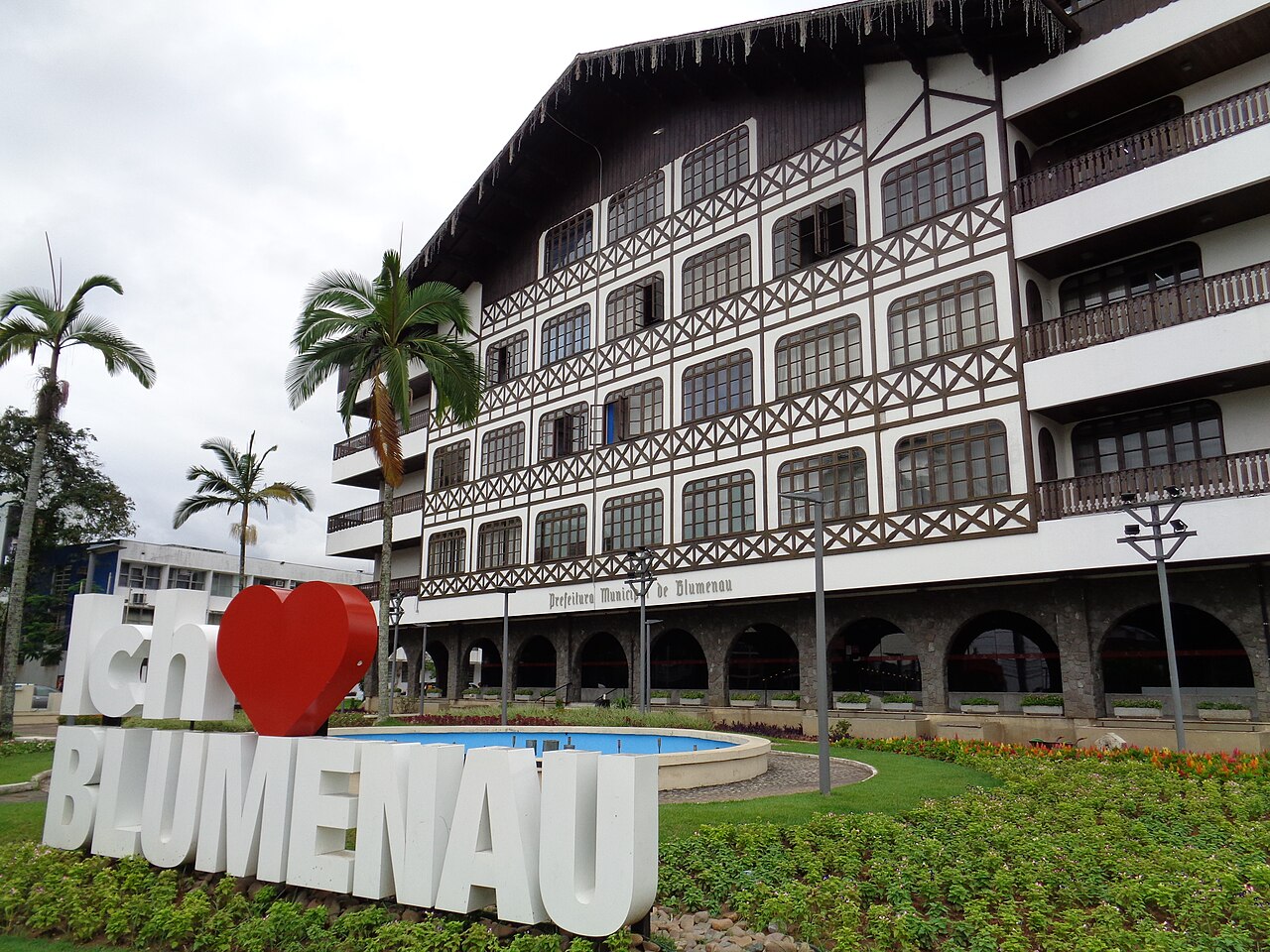 Letreiro com Ich, coração vermelho e Blumenau diante da Prefeitura, com fachada branca, estrutura escura, palmeiras e espelho-d’água.