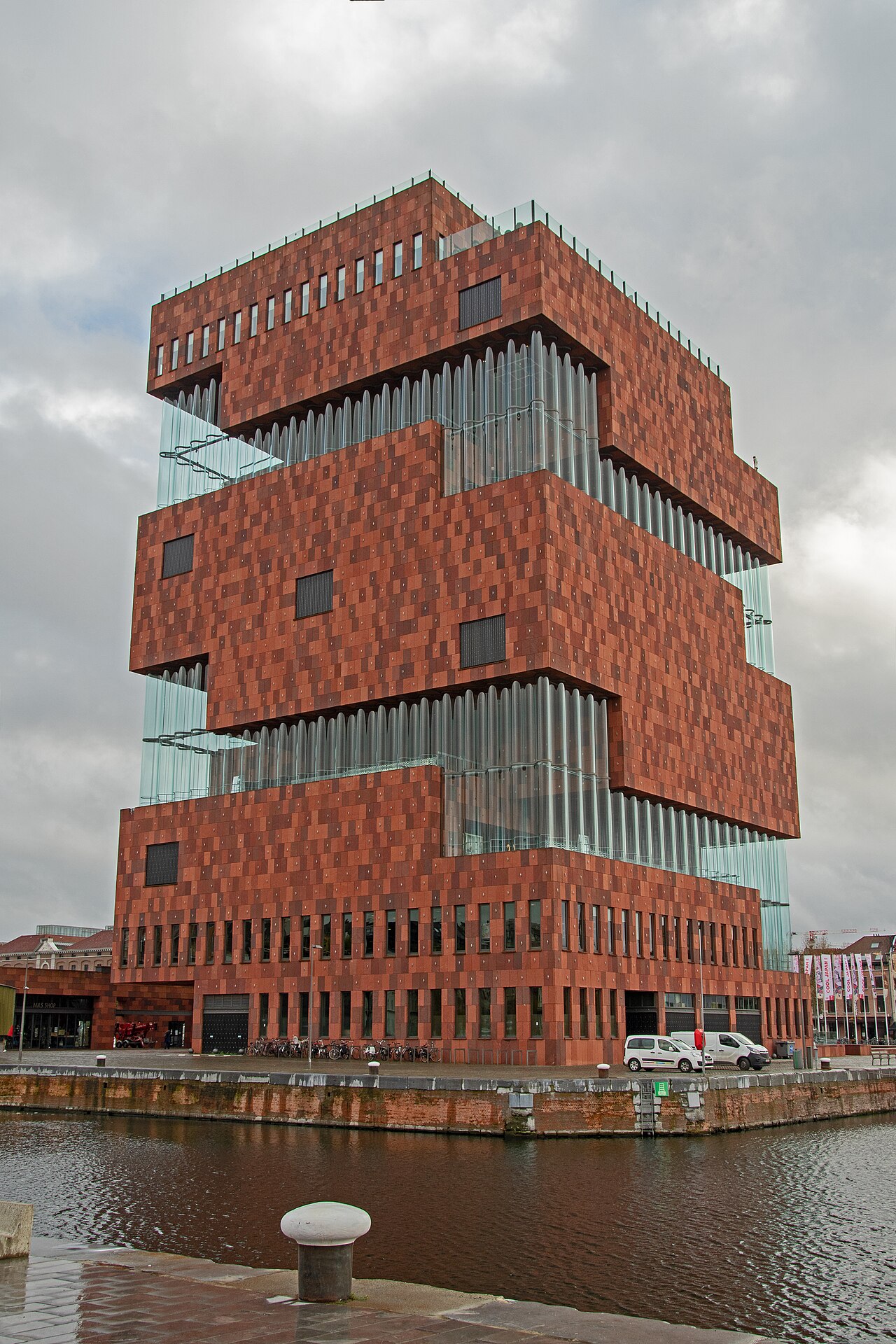 Edifício avermelhado do MAS com volumes sobrepostos e faixas de vidro ondulado, visto diante de uma doca sob céu encoberto.