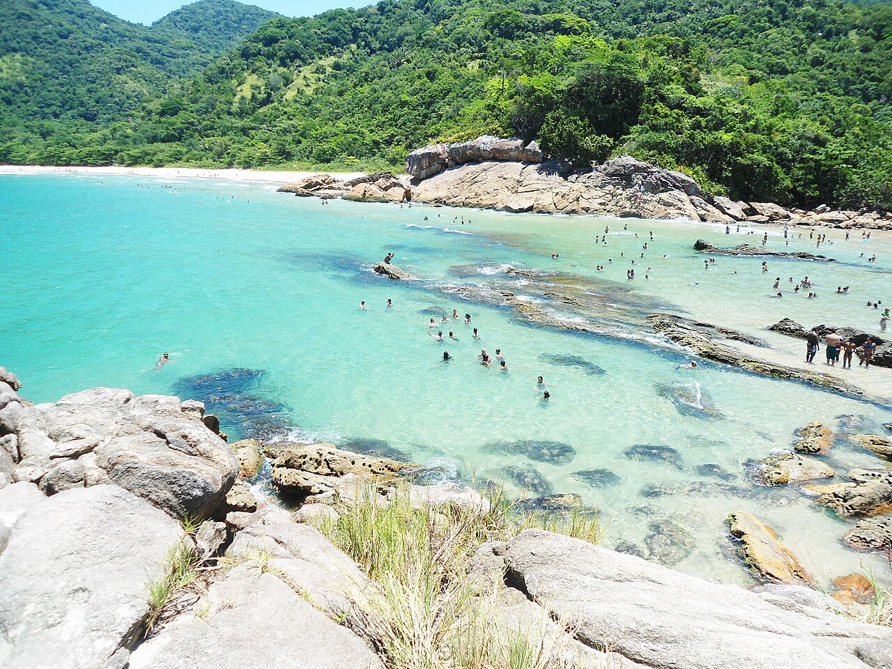 Água azul-turquesa com banhistas entre rochas na Praia do Meio, faixa de areia ao fundo e morros cobertos por vegetação.