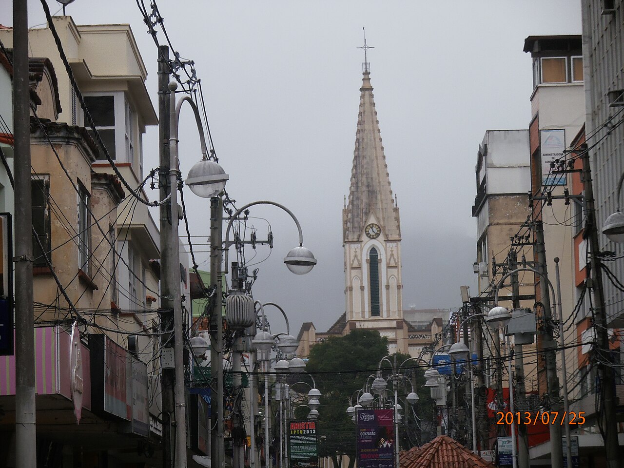 Rua entre prédios comerciais, com fios e postes, e a torre da Igreja de Santa Teresa ao fundo sob céu encoberto.