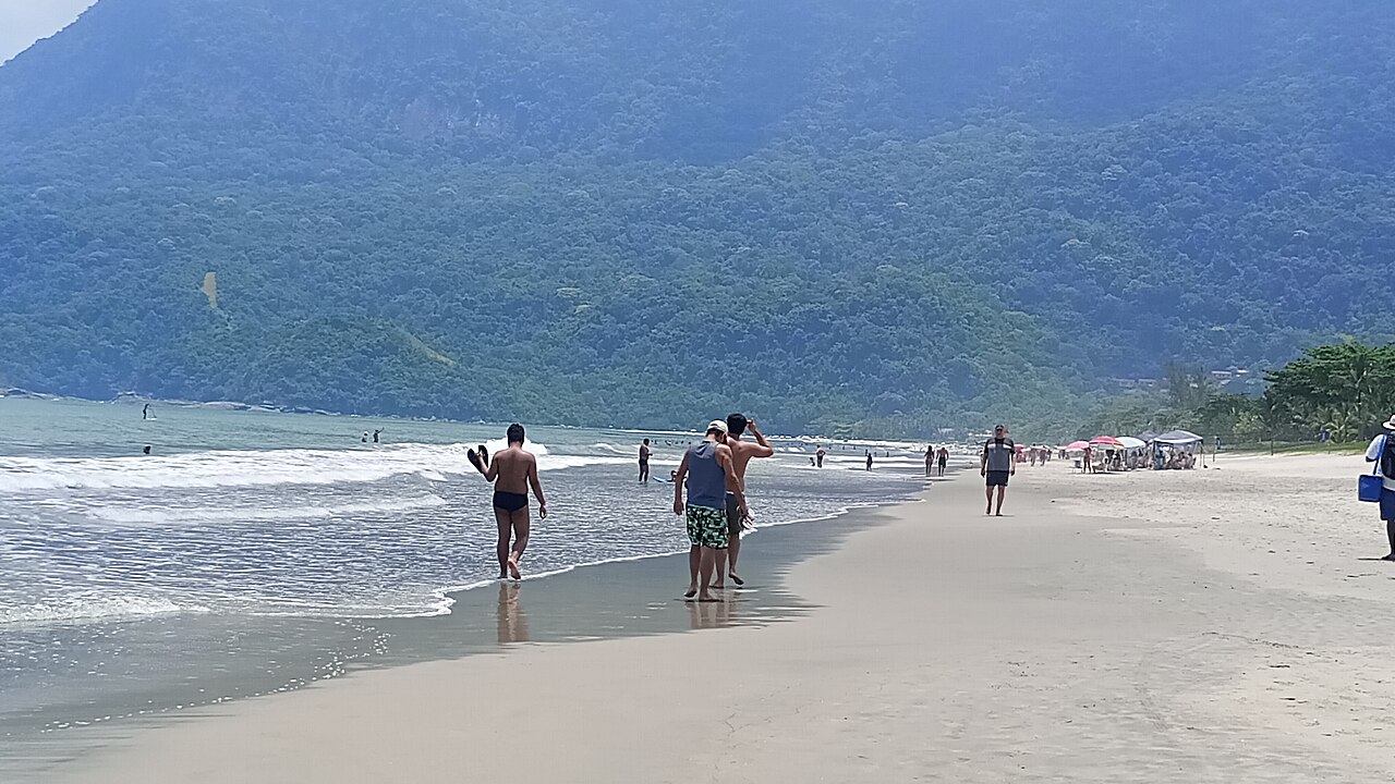 Faixa de areia em Guaecá com pequenas ondas, banhistas, pessoas caminhando, guarda-sóis e um morro coberto de vegetação ao fundo.