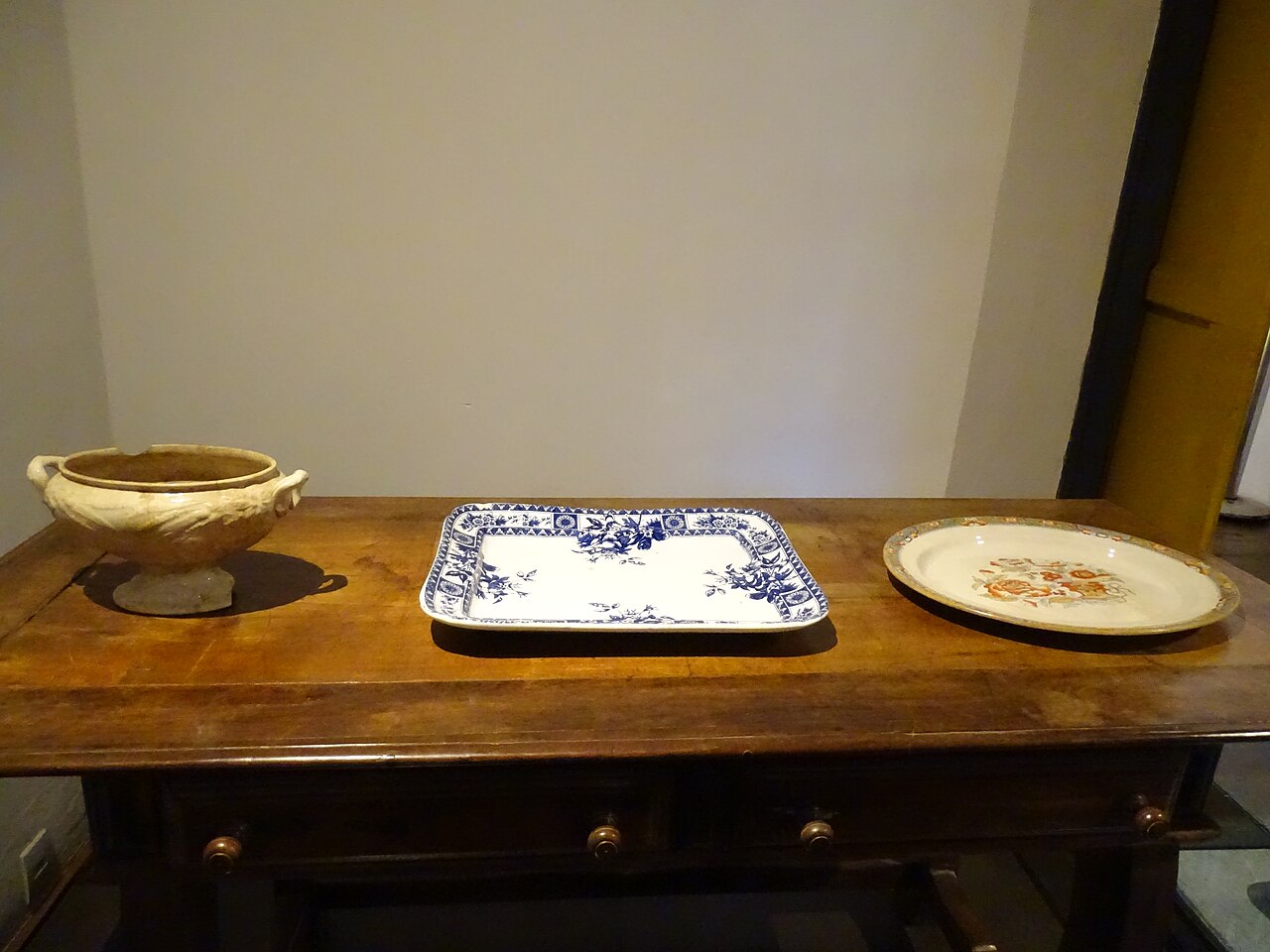 Móvel de madeira escura com três peças de louça em exposição no Museu Casa Padre Toledo: um recipiente com alças e duas travessas decoradas.